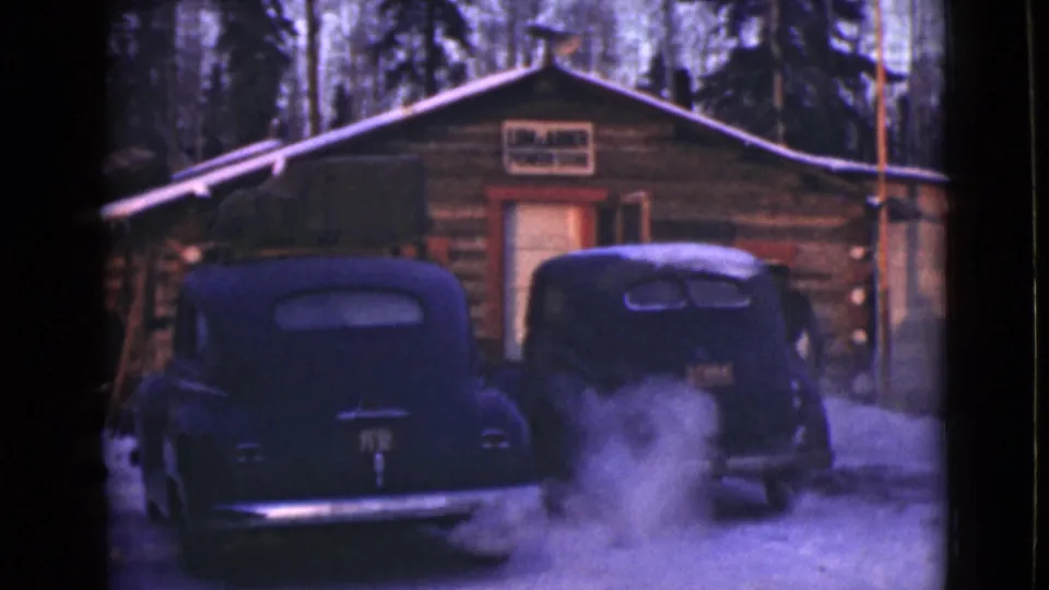 Snowy Alaska cabin scene with two parked black cars and visible steam or exhaust rising near the right car.