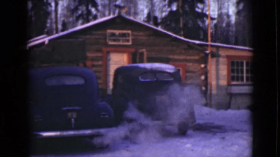 1946 Alaska home-movie frame with black cars close to camera, steam in the snow, and the cabin entrance behind them.