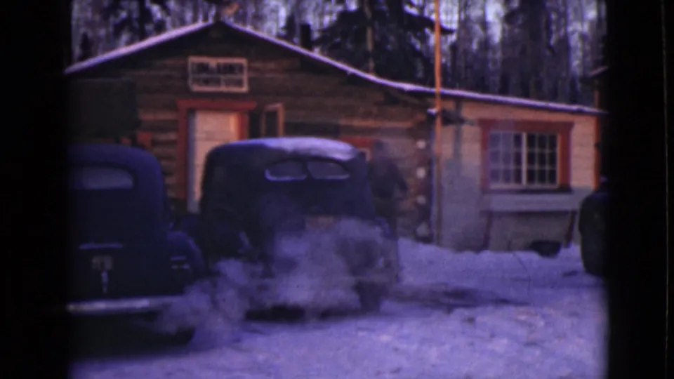 Snowy 1946 Alaska cabin view with a steaming vintage car and an indistinct person partly visible near the right edge.