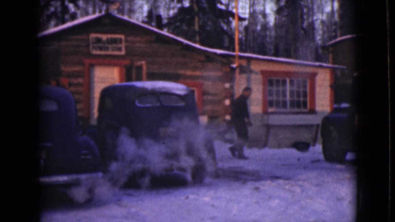 1946 Alaska home-movie frame showing a snowy log cabin, parked old cars, rising exhaust, and a man near the doorway area.