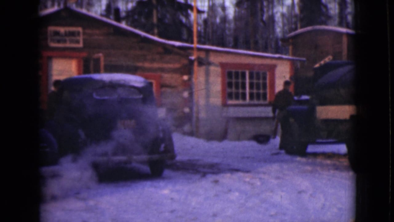 Snowy cabin yard with a man near an old vehicle and exhaust drifting in the cold air.