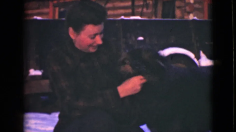 A woman in a dark winter coat gently touches a large dog in a snowy Alaska setting, with 8mm frame borders visible.