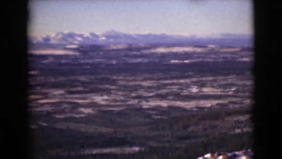 1946 8mm Alaska home-movie frame showing a distant snowy mountain range beyond a wide valley and forested hills.