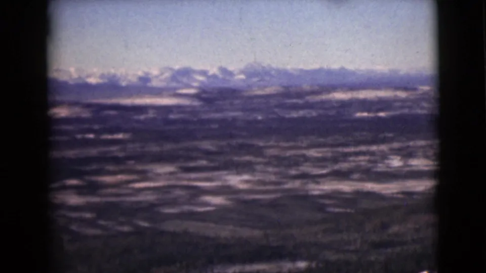 1946 Alaska 8mm frame with snowy peaks on the horizon and layered valley terrain below a pale blue sky.