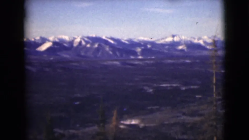 1946 Alaska 8mm frame showing a rugged snow-capped ridge line, open sky, dark valley, and thin foreground trees from a scenic overlook view.