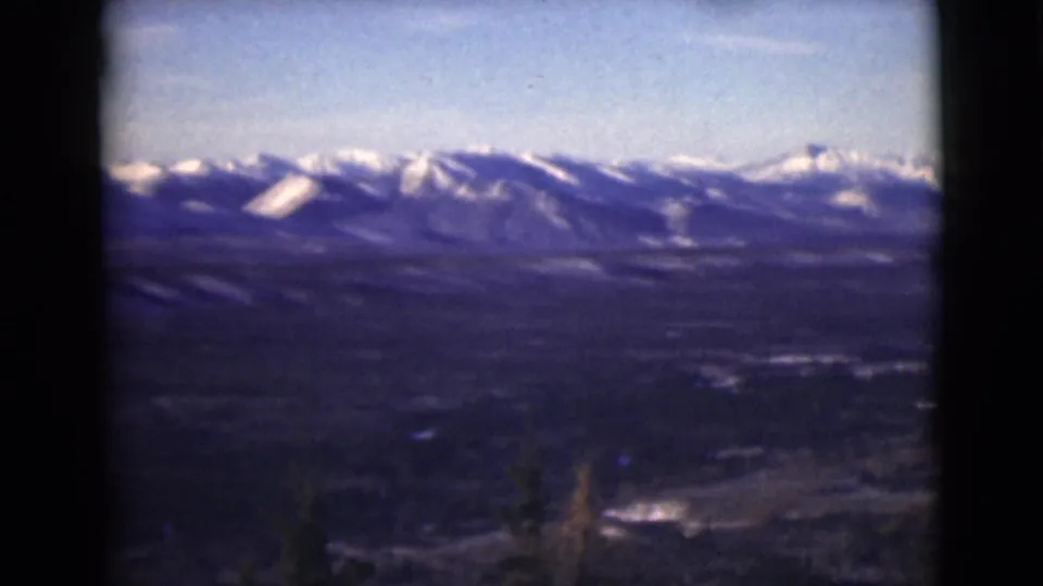 1946 Alaska 8mm frame showing a distant snowy mountain range, broad valley, blue sky, and dark evergreen foreground.