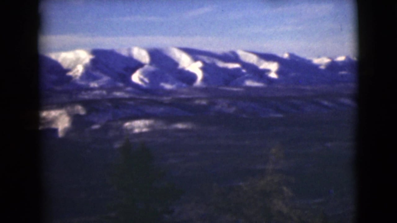 1946 Alaska 8mm frame showing distant snow-capped mountains beyond a dark, forested foreground slope.
