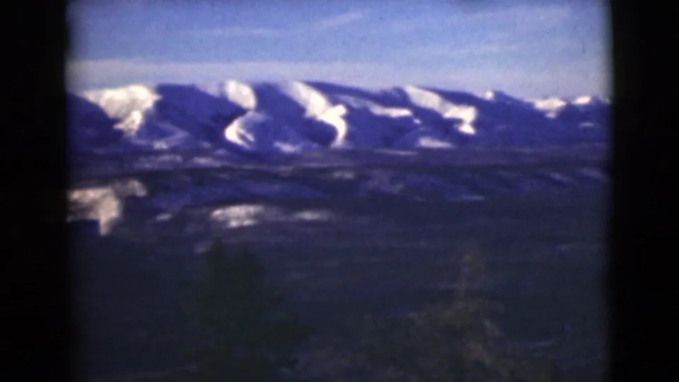 1946 Alaska 8mm frame showing distant snow-capped mountains beyond a dark, forested foreground slope.