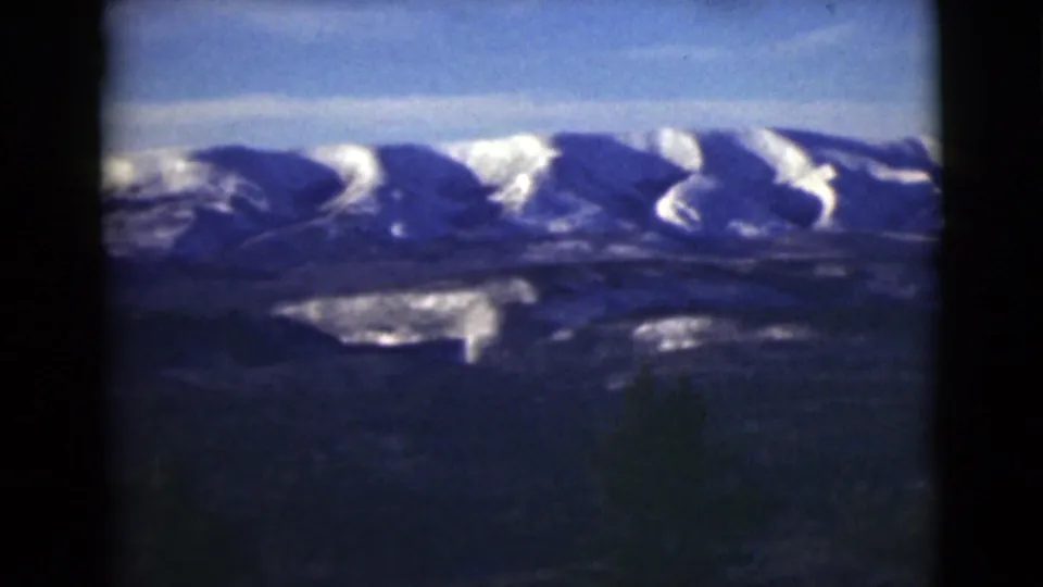 Vintage 8mm Alaska frame with snow-covered peaks above dark lower terrain and a pale blue sky.