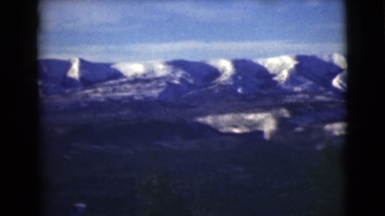 Snow-covered mountain range under blue sky with dark foreground slopes and film borders.