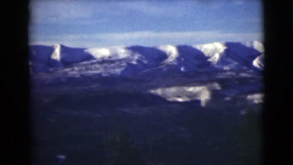 1946 Alaska 8mm frame showing distant snowy mountains, shadowed terrain, and a blue sky from a high vantage point.