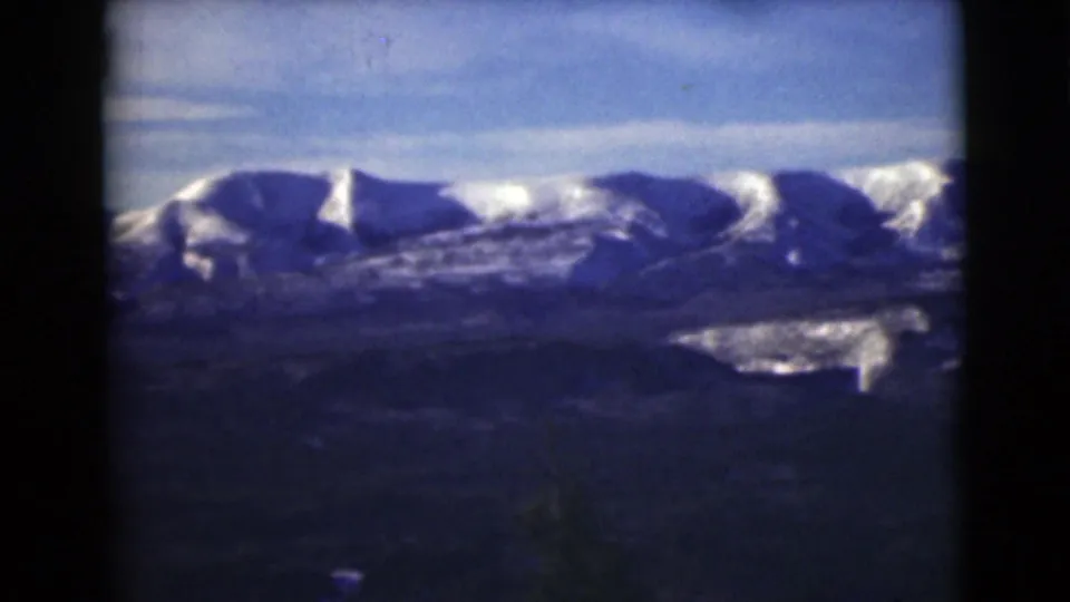 1946 Alaska home-movie frame with a panoramic view of snowy ridges, dark slopes, and soft 8mm color texture.
