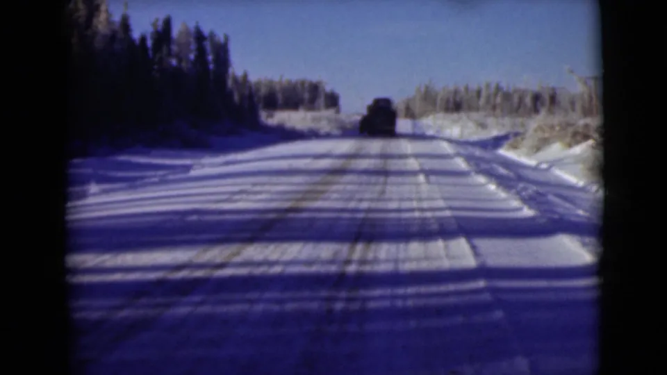 1946 Alaska 8mm frame looking down a snow-packed road bordered by frozen forest, with a dark vehicle ahead.