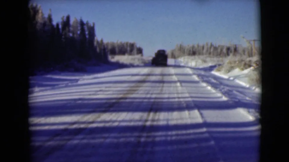 1946 Alaska 8mm frame showing a snow-packed forest road receding toward a dark vehicle under clear sky.