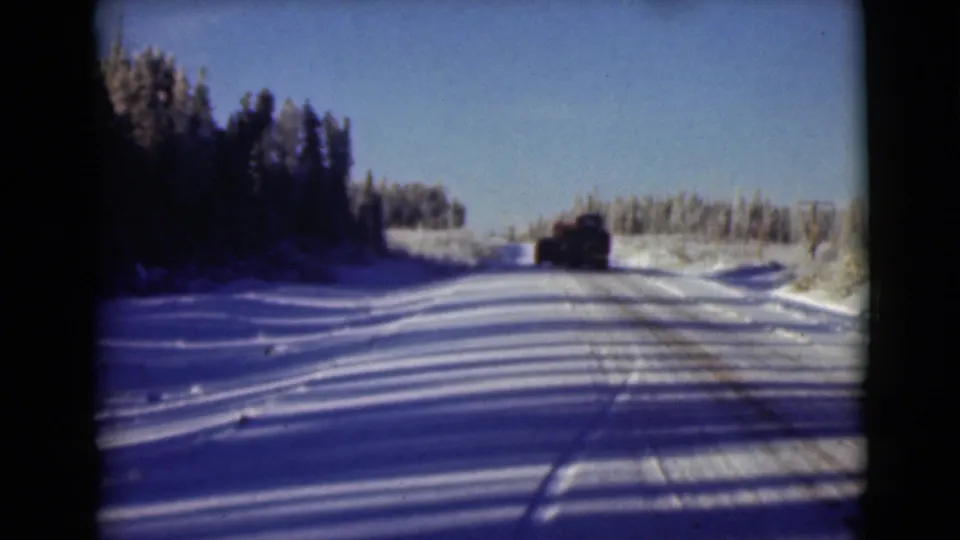Vintage 8mm Alaska frame with a lone vehicle receding along a snowy forest road.