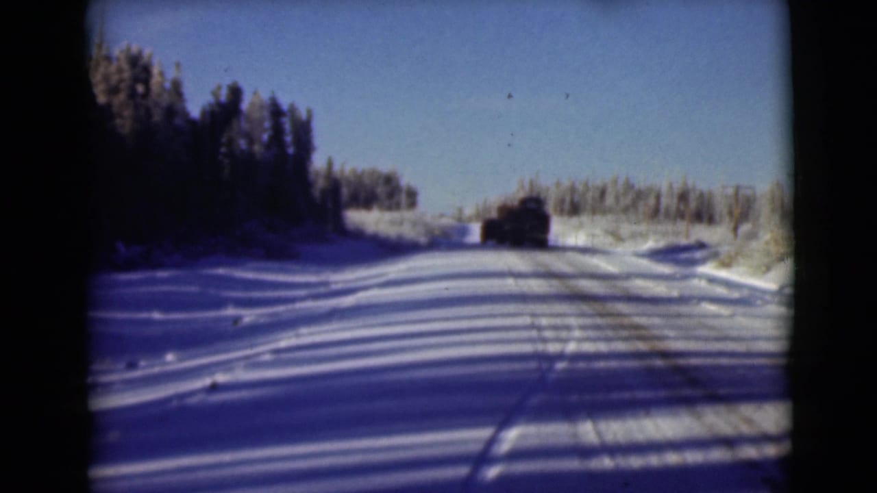 Snowy road with tire tracks, evergreen trees, and a dark vehicle driving away.