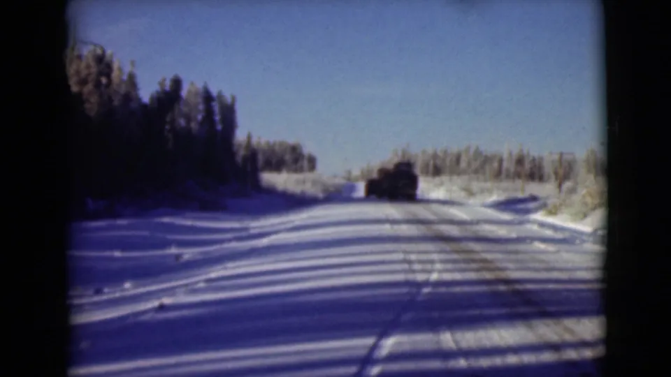 1946 Alaska home-movie frame with a vehicle ahead on a snowy forest road under clear blue sky.