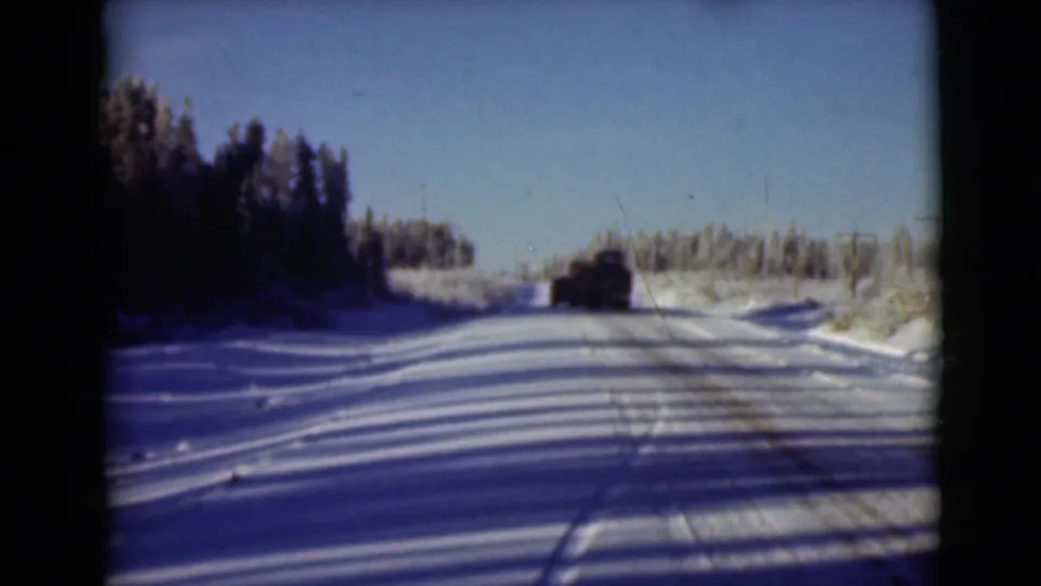1946 Alaska home-movie frame of a vehicle receding along a snowy road through evergreen forest.
