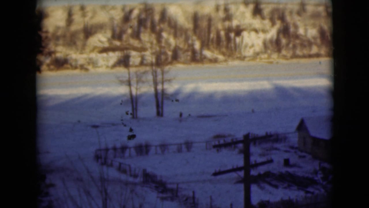 Snowy field with bare trees, distant wooded hills, fencing, a small structure, and a utility pole.