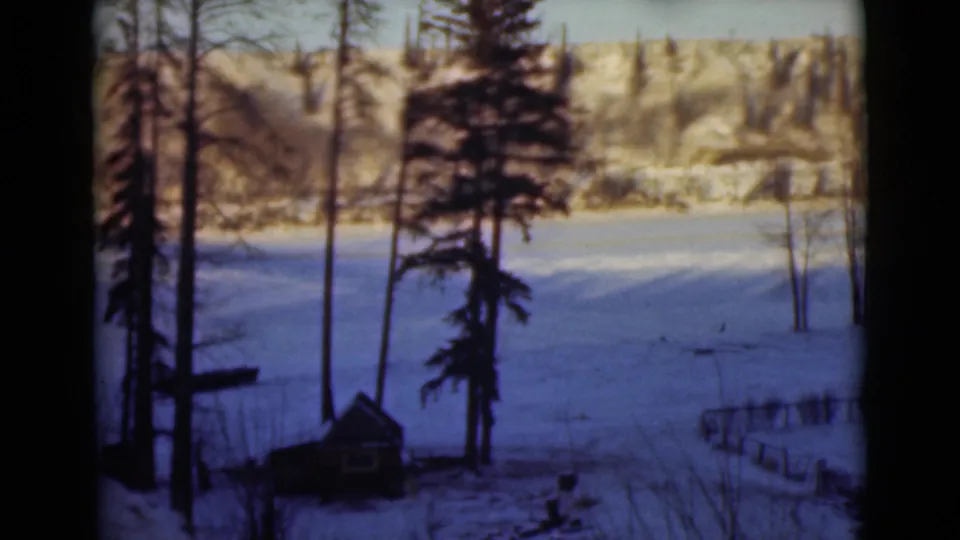1946 Alaska home-movie frame of a snowy field framed by trees, with a small structure, low fencing, and wooded hills beyond.