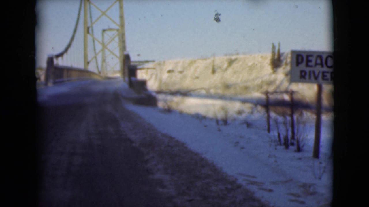 1946 8mm frame of a snowy road leading toward a steel suspension bridge, with a roadside sign reading Peace River.