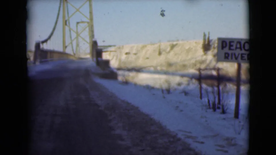 1946 8mm frame of a snowy road leading toward a steel suspension bridge, with a roadside sign reading Peace River.