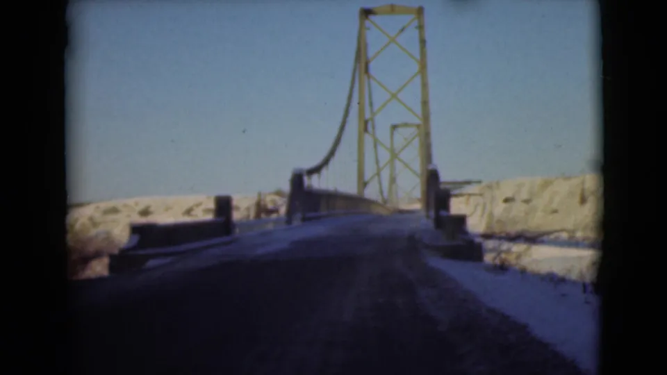 1946 8mm frame showing a forward road view toward yellow steel bridge towers in a snowy Alaskan landscape.