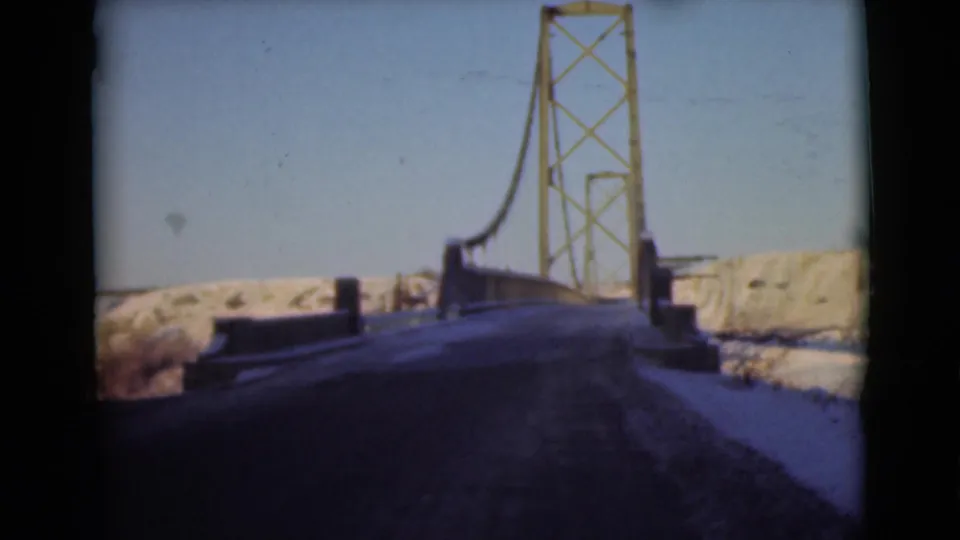 1946 8mm frame from a moving vehicle nearing a suspension bridge over a snow-covered northern landscape.