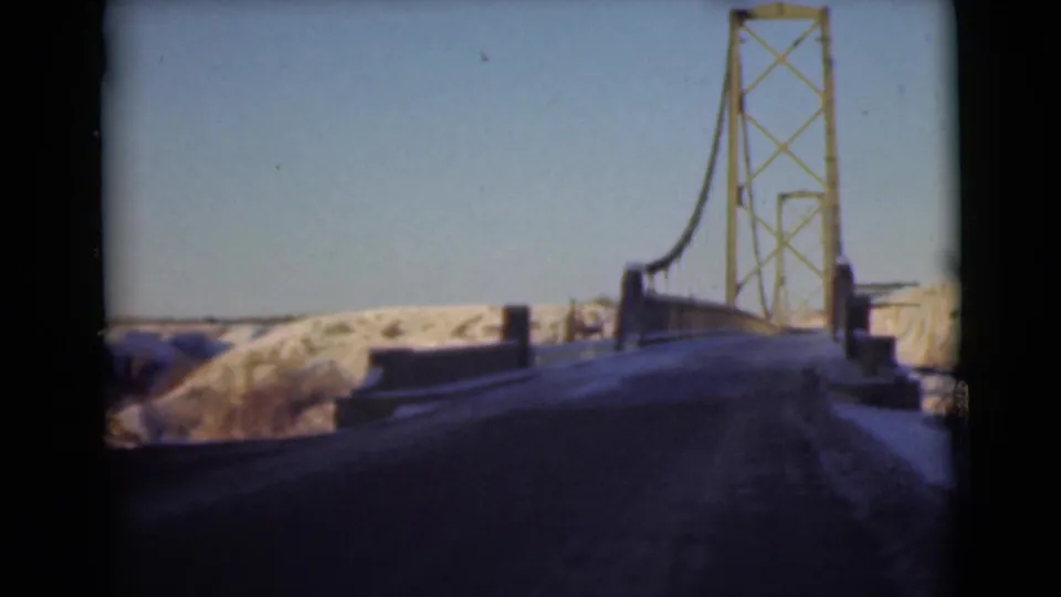 1946 8mm frame of a snow-lined road at the approach to a yellow suspension bridge in the Alaska footage.