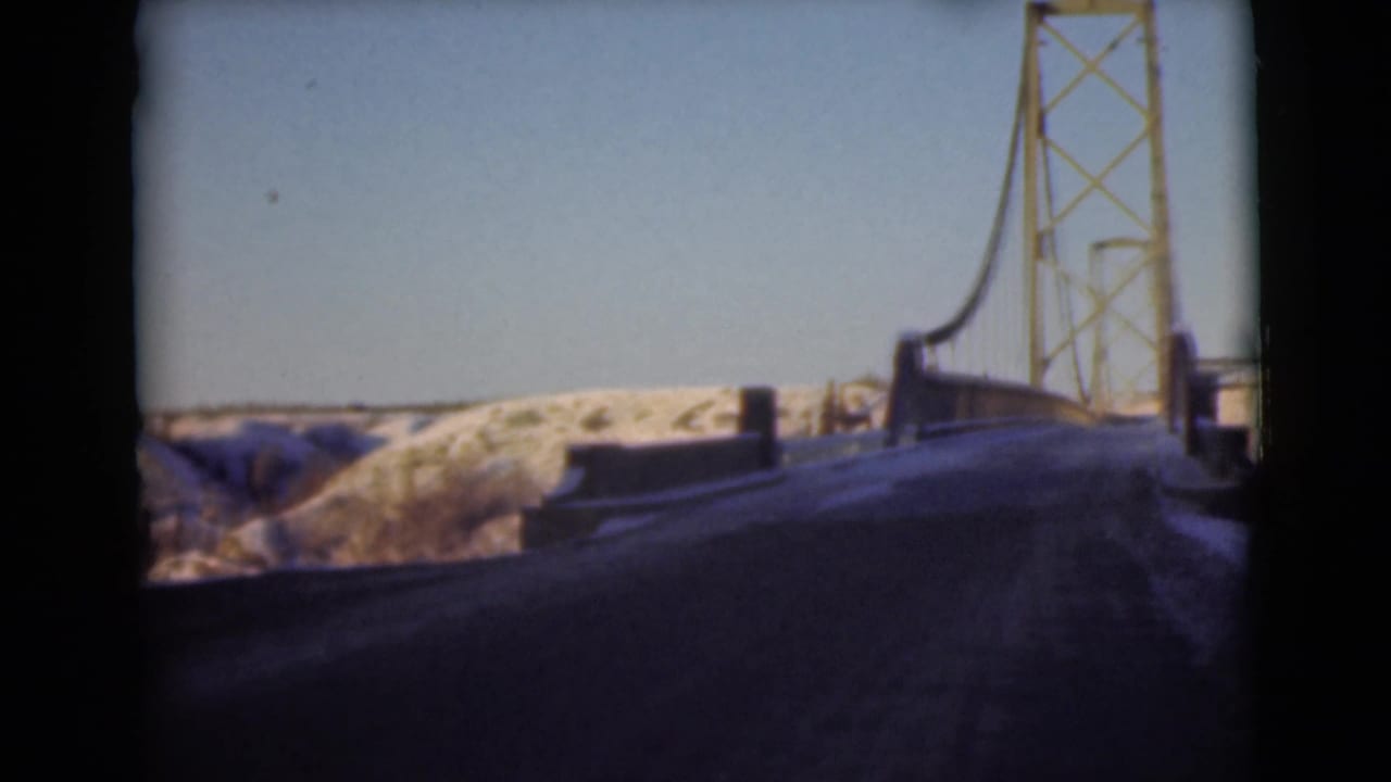 Roadway approaching a suspension bridge with snowy hills beyond.