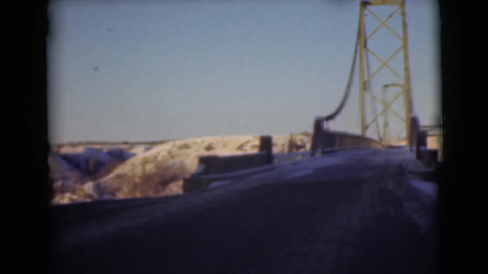 1946 Alaska 8mm frame showing a dark road leading toward a suspension bridge tower with snow-covered hills in the distance.