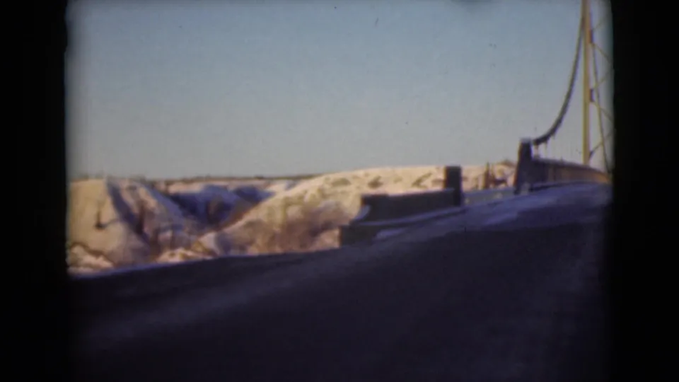 1946 Alaska travel footage frame with a bridge edge at right, dark road surface, and snow-covered hills under a pale blue sky.