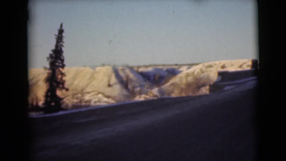 1946 Alaska frame with a lone evergreen near the bridge roadway, snow-covered hills, and clear sky in the background.