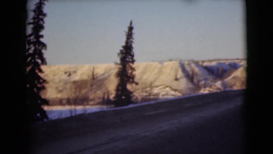 1946 Alaska 8mm view of a dark road bordered by snow, evergreen trees, and low winter hills.