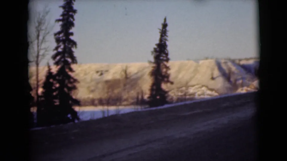 1946 Alaska frame with roadside evergreens, a bare tree, snow patches, and winter hills beyond a dark road.