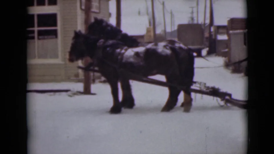 1946 Alaska frame of two dark horses hitched to a wooden sled on a snowy town street near simple buildings.