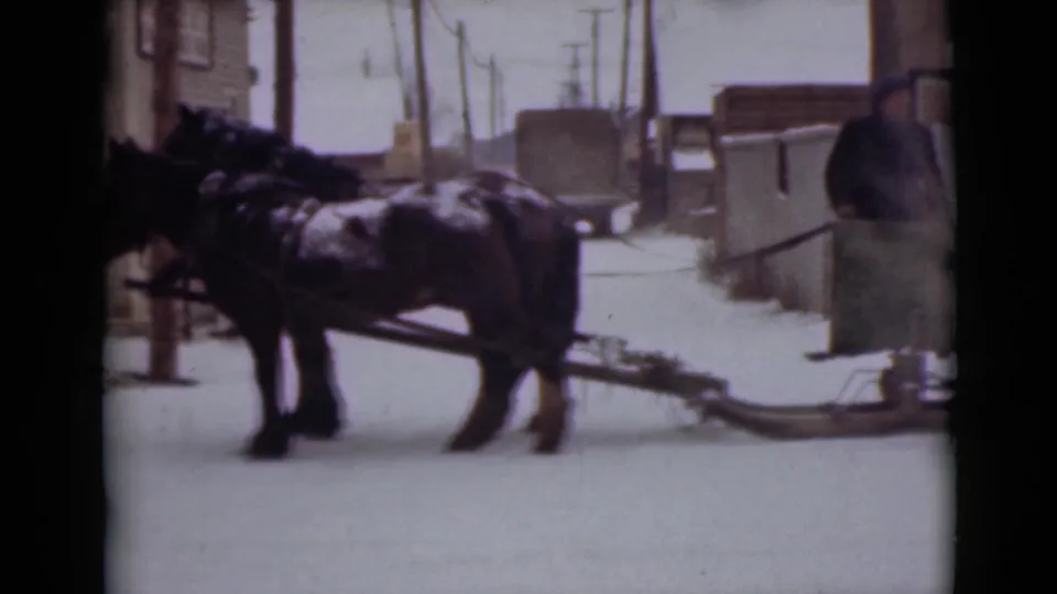 1946 Alaska home-movie frame showing harnessed horses and a low sled in a snowy town setting with utility poles.
