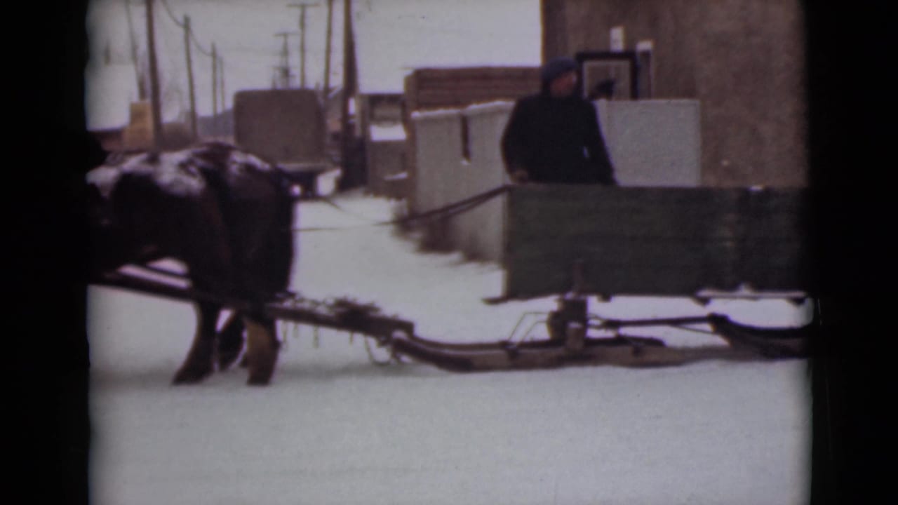 Horse-drawn sleigh moving along a snowy street beside town buildings.