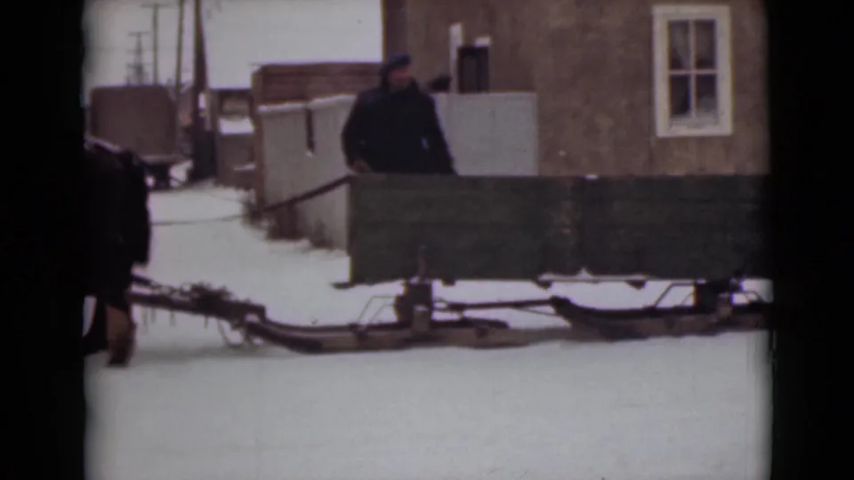 A horse-drawn green sleigh continues past a plain building as a bundled adult man stands in the snowy Alaska street scene.