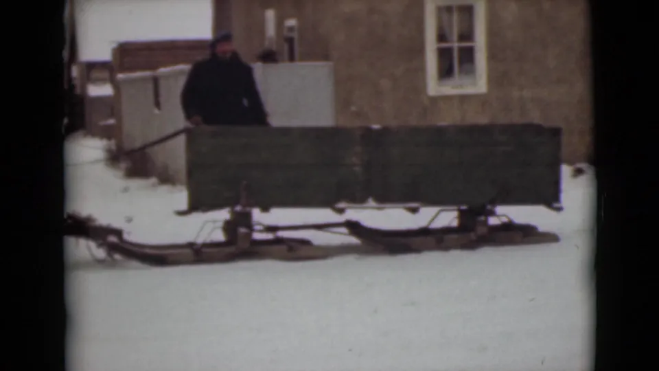The 8mm frame shows a bundled man standing near the green sleigh with snow underfoot and a simple town building behind him.