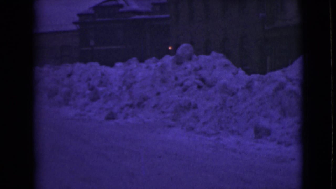 Purple-tinted 8mm frame of high snow drifts along a quiet road beside buildings.