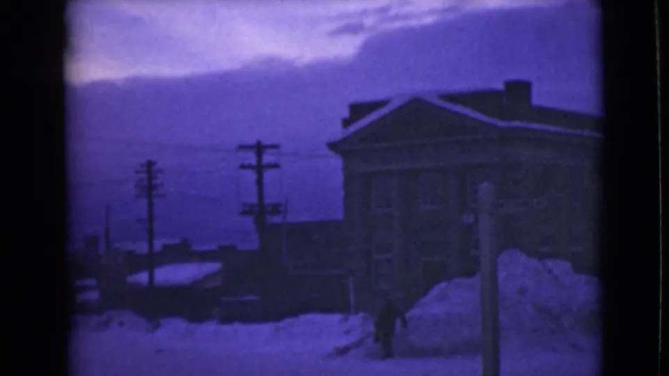 1946 Alaska 8mm frame with a lone distant figure walking by snowbanks, utility poles, and town buildings.