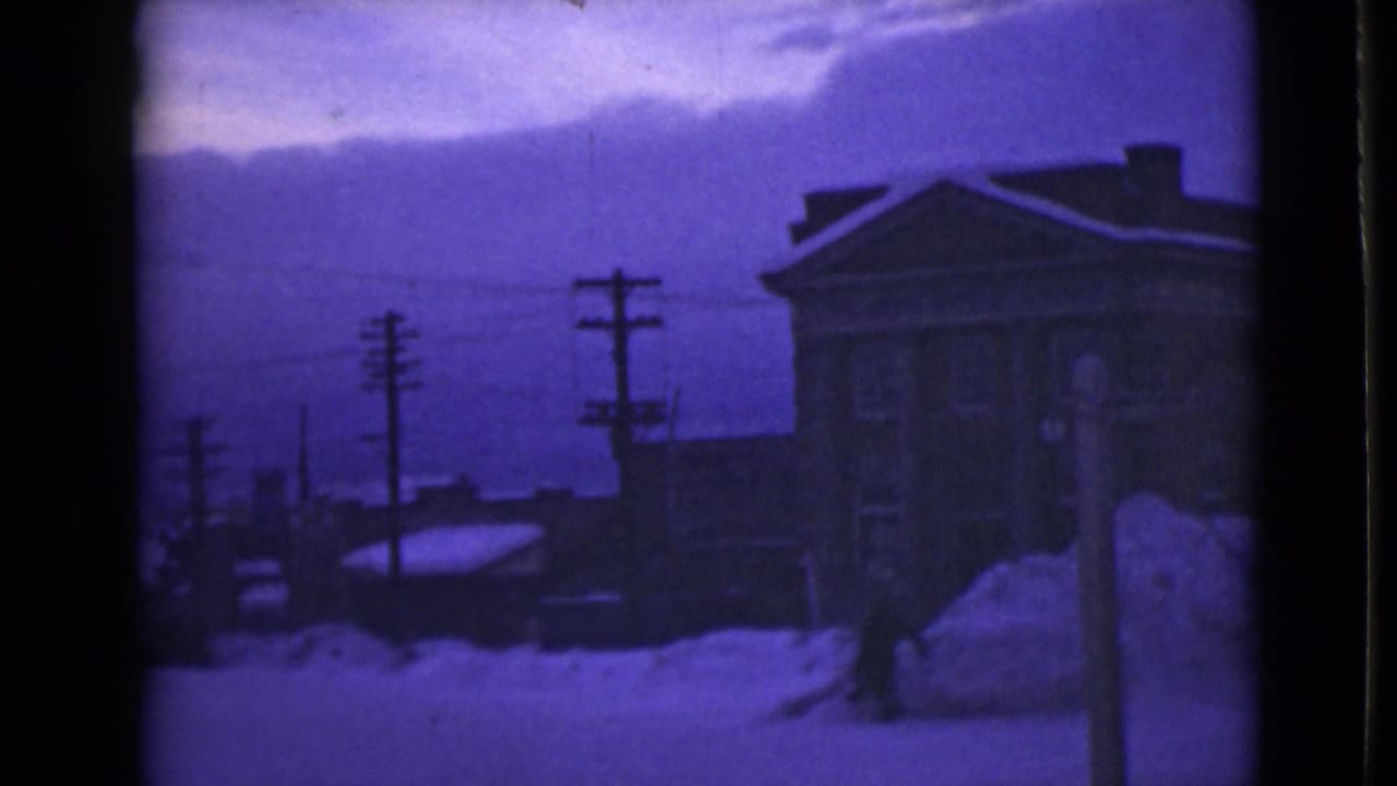 Snow-covered street with tall drifts, utility poles, a large building, and a distant figure near the roadside.