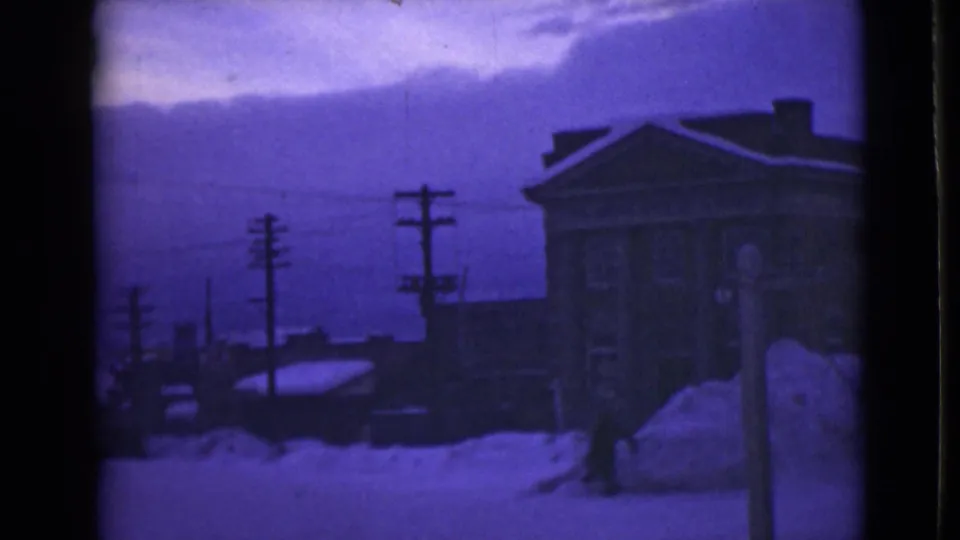 1946 Alaska home-movie frame showing a snowbound street, tall roadside drifts, utility poles, a large building, and a distant person near the snowbank.