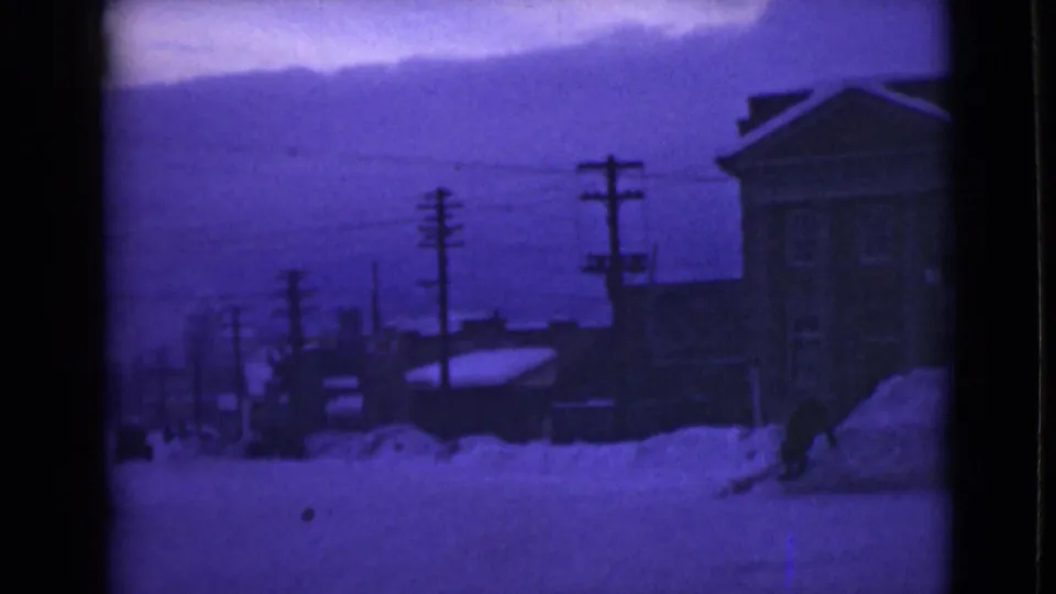 1946 Alaska frame with a snow-covered road, high drifts, utility poles, rooftops, and a large building along the right edge.