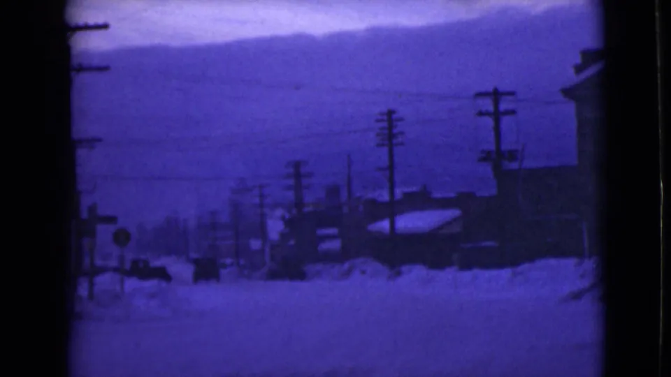 1946 Alaska frame showing a quiet snowbound street, utility poles, heavy roadside drifts, a large building, and distant hills in purple-tinted 8mm film.