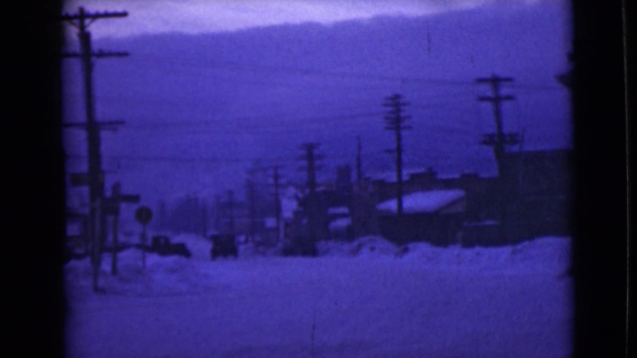 1946 Alaska home-movie frame showing a snow-covered street, utility poles, low buildings, and a distant car under blue color shift.