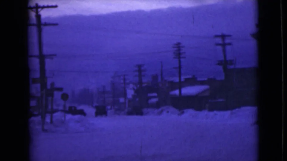1946 Alaska home-movie frame showing a snow-covered street, utility poles, low buildings, and a distant car under blue color shift.