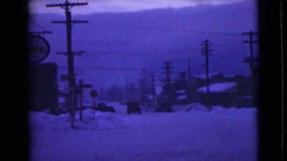 1946 Alaska street view with snowbanks, utility poles, a slow car, low buildings, and a partially visible Imperial sign.
