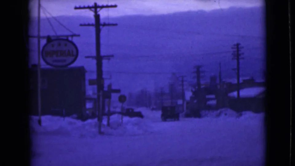 1946 Alaska home-movie frame showing an Imperial sign beside a snowy street with poles, wires, buildings, and a car ahead.